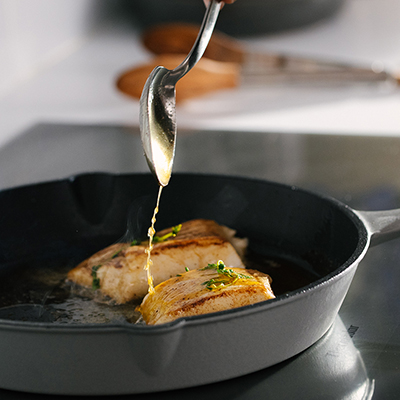 Pan fried sea bass in a cast-iron with a spoon dripping butter on top.