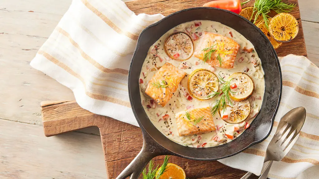 Cast iron skillet full of salmon, a creamy sauce, and slices of lemon.