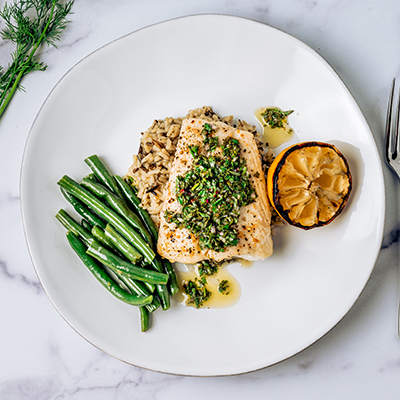 Top view of halibut fish with chimichurri sauce, green beans, lemon on a white plate on marble table