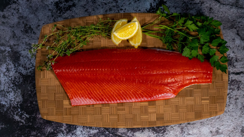 Copper river king salmon season with a filet of salmon on a wooden block surrounded by herb garnishes.