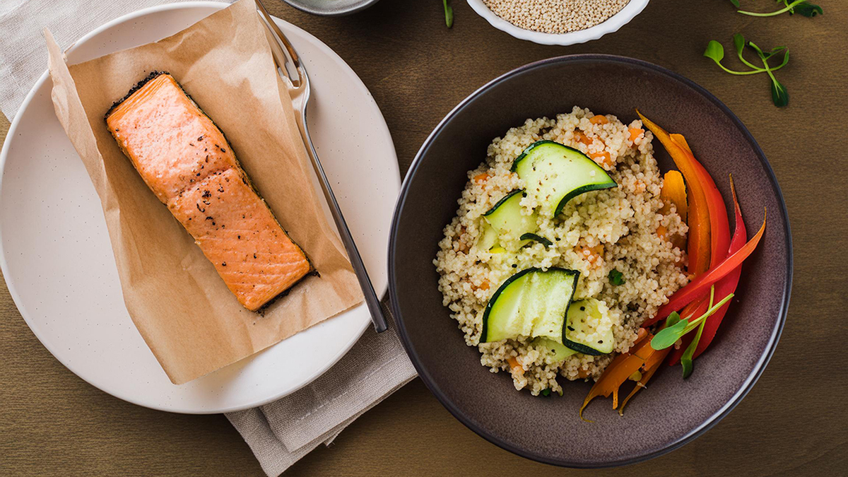 Mediterranean menu with a piece of cooked salmon in parchment paper next to a bowl of quinoa and vegetables.